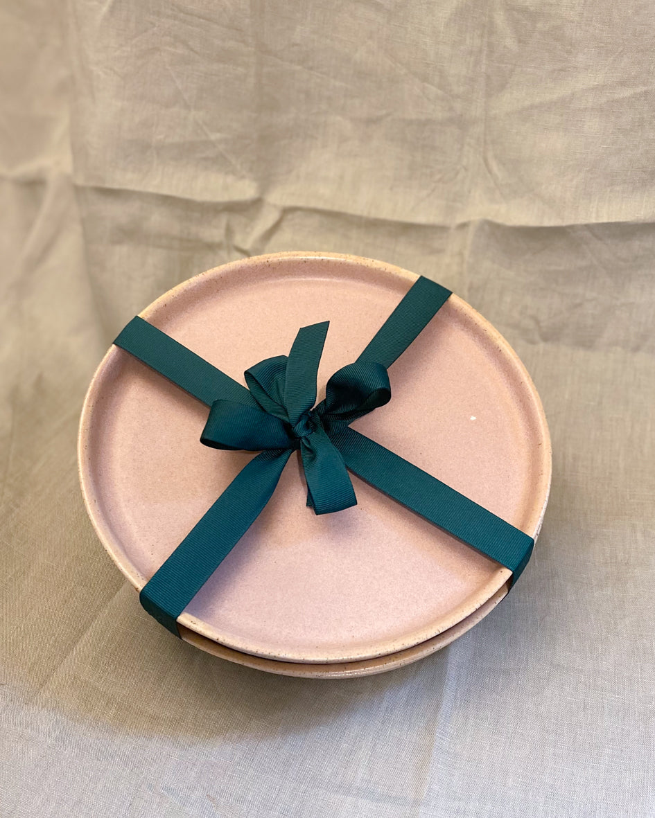 A stack of blush pink plates seen from above. A dark green ribbon is tied around the stack with a bow.